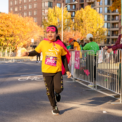 Girls on the Run participant wearing a pink cape and mask crossing the 5K finish line.